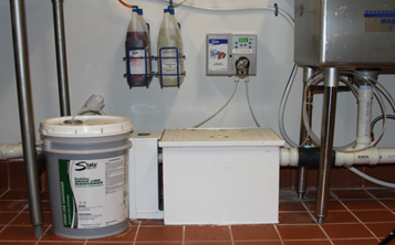 A gallon bucket of a State Chemical branded bucket is pictured next to a white rectangular box. To the right of the box, there is the bottom of a sink. The State branded box is hooked up to the sink through a clear tube. The tube slowly dispenses product from the bucket to the sink drain.